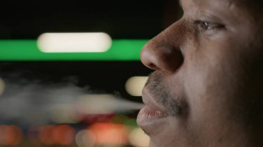 Black teenager smokes cigarette with nicotine. Close-up face African American smoking cigarette with marijuana, exhales smoke. Bad cigarette smoking habit. Concept smoking cannabis, cigarette smoking