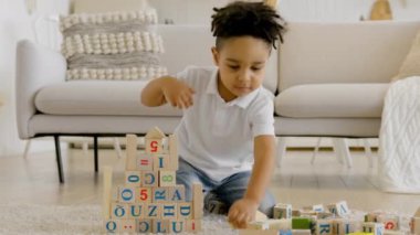 Child builds tower of wooden cubes at home. Happy African American cute little boy is playing game, building tower of colorful wooden blocks, sitting on floor in bright living room of house