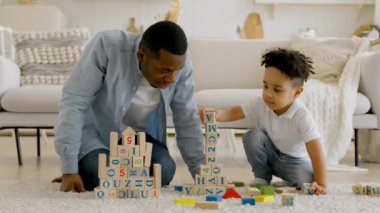 Happy cute little son is playing game with black dad, babysitter, tower builder made of colorful wooden blocks, father of an African family and little boy are having fun on warm floor of house