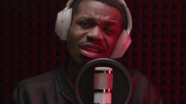 Close up portrait young African American handsome and stylish singer gesturing with his hands, recording song and singing into microphone in sound studio headphones on red studio lights