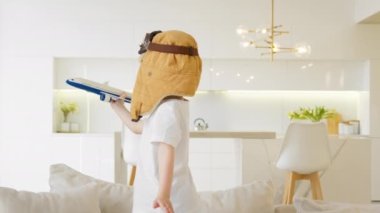 Happy child is spinning around playing with modern toy plane at home, flying up and down holding plane in his hands in white t-shirt and brown hat on his head. Eagerness to fly up on air
