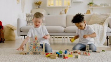Two happy 4 year olds boys of different ethnic groups play together building towers and pyramids from wooden blocks sitting on floor of living room. Little children, childrens friendship