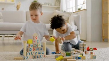 Childrens friendship two happy 4 year olds boys of different ethnic groups play together building towers and pyramids from wooden blocks sitting on floor of living room. Little children