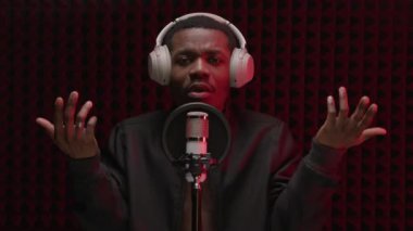 Rap singer singer sings in microphone. African American singer performs song while standing in recording studio with headphones on his head and sings into microphone, emotionally waving his arms