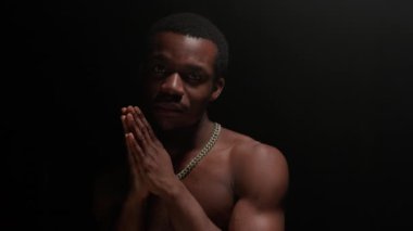 Portrait serious African American man experiencing onset of tragedy in life and praying with his hands clasped in front of him with clasped praying hands in dark studio with one light source on top