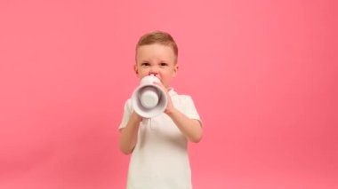 Portrait 5 years boy in white T-shirt shouts expresses his dissatisfaction, swears masher menacingly shouts into megaphone on pink background. Kid shouts emotionally into loudspeaker waving his hand