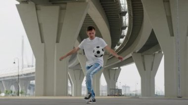 Professional football player professionally owns juggles ball in city. Caucasian teenager man in white T-shirt and blue jeans football freestyler juggles soccer ball, shows various tricks with ball