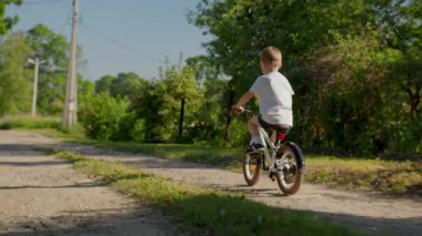 Child riding bicycle on dirt road in country village in summer sunny weather. Child rides bicycle in summer weather with toy multi-colored windmill. shooting from behind