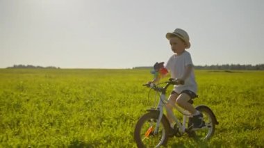 Happy blond 4-year-old boy road across a field of green grass on white bicycle with rays of setting sun with toy windmill toy. Child enjoys a carefree childhood and freedom riding bicycle