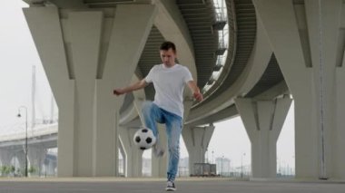 Man soccer player is practicing kicks tricks with ball in city under the bridge improving his skills. Footballer training outdoors juggling ball. Wearing casual style. Sports football skills concept.