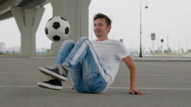 Young freestyler wearing blue jeans and black sneakers juggles ball sitting on empty roadway. Man smiles doing tricks. Sportsman enjoys leisure activity.