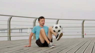Young sportsman does air move tricks sitting on path. Professional freestyler juggles ball using feet. Young man demonstrates powerful juggling skills
