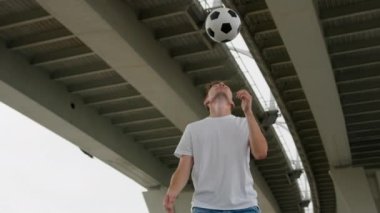 Stylish sportsman juggles football on head under overpass highway. Young man wearing white shirt throws ball and demonstrates trick doing head bounce.