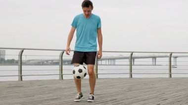 Professional sportsman juggles football on wooden bridge. Young man shows tutorial of balancing ball against grey river and sky. Guy enjoys freestyle football