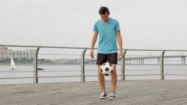 Young sportsman does ball freestyle tricks on bridge. Stylish man wearing blue t-shirt and black shorts juggles and holds football on knees against grey river and sky