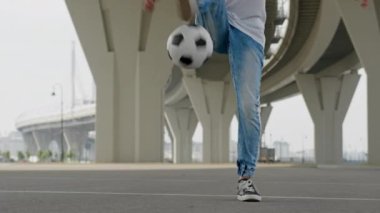 Stylish man wearing blue jeans shows tricks of balancing and kicking ball on feet. Young sportsman demonstrates professional juggling ball under city overpass