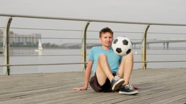 Young freestyler juggles ball sitting on pathway against boat on river. Sportsman does air move tricks. Man enjoys being professional freestyler at sunlight