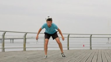 Young man twists ball on shoulders doing large circles on empty path. Sportsman juggles ball on upper body. Man shows perfect skills against blurry grey bridge