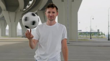 Young man wearing white t-shirt juggles ball using finger under trestle. Adult enjoys life being professional freestyler. Sportsman smiles and looks straight closeup
