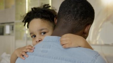 Multiracial boy embraces a black African-American father and looks into camera. Hugs of father and 3-year-old son sitting on couch at home. Happy multiracial family of father and son