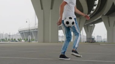 Young sportsman wearing blue jeans juggles ball backwards using heels on empty road. Stylish man demonstrating tricks backwards under trestle slow motion