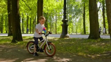 Happy blonde smiling child is having fun riding around park on white bicycle with toy multi-colored windmill among green trees in sunny summer weather in park. Carefree freedom childhood of child