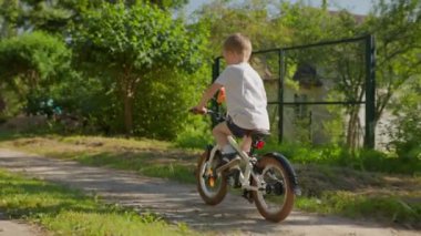 Child rides bicycle in summer weather with toy multi-colored windmill. shooting from behind, slow motion video. Child riding bicycle on dirt road in country village in summer sunny weather