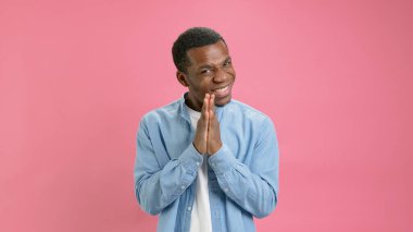 Black man asks for forgiveness by folding his hands together on pink background. Young African American man looks hopefully into camera, puts his hands in front of him and asks, begs to forgive him
