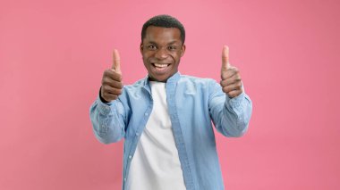 Kendine güvenen lider, kot gömlek ve beyaz tişört giyen genç Afro-Amerikalı, basit pembe stüdyo arka planında izole edilmiş bir jest olarak el hareketi yapıyor.