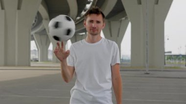 Young man wearing white t-shirt demonstrates football trick under trestle. Freestyler twists ball only on finger and looks straight closeup. Guy enjoys life
