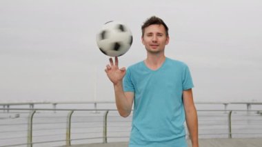 Young man wearing blue t-shirt twists ball on finger. Stylish sportsman looks straight juggling football. Professional sportsman enjoys life being freestyler