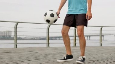 Young man does air move tricks on wooden embankment. Stylish sportsman wearing blue t-shirt and black shorts juggles football against grey river with bridge and sky