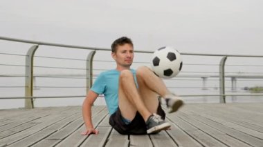 Young man in blue t-shirt does sitting tricks on boardwalk. Sportsman juggles ball with feet. Guy shows tutorial of juggling with smile. Man enjoys being freestyler
