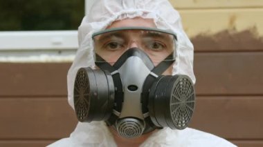 Brown-eyed man stands against painted wooden house wall. Workman equipped with special protective clothes and respirator with goggles to avoid poisoning closeup