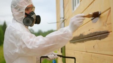 Adult man paints wooden house outer board wall with brown paint. Workman in respirator with goggles and special overall holds bucket with paint closeup