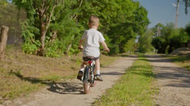 Child riding bicycle on dirt road in country village in summer sunny weather. Child rides bicycle in summer weather with toy multi-colored windmill. shooting from behind, slow motion video