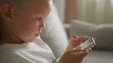 Little boy dressed in white T-shirt sits on sofa with smartphone in hands. Concentrated preschooler flips through photo gallery on touch-screen phone closeup