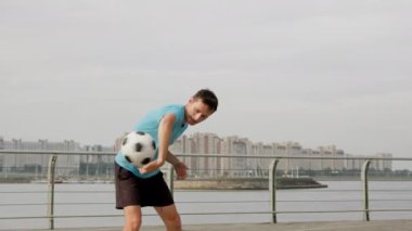 Young sportsman balances ball against city buildings. Freestyler wearing blue t-shirt twists ball using shoulders and fingers. Man demonstrates skills on juggling