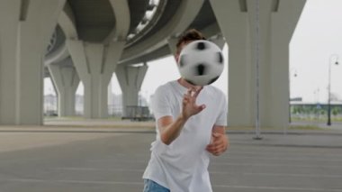 Young sportsman throws ball and twists on finger on nasty day. Stylish guy demonstrates professional skills on juggling football under trestle closeup