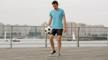 Young freestyler wearing blue t-shirt and black shorts juggles ball on wooden bridge. Stylish sportsman does air move tricks balancing ball against city buildings