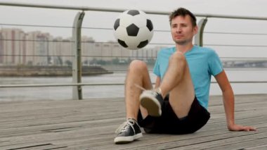 Young man does sitting tricks on boardwalk. Sportsman in blue t-shirt and black shorts juggles ball sitting on pathway. Guy enjoys life being freestyler against sky
