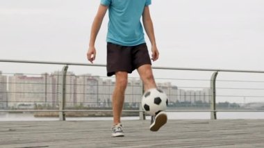 Young man juggles ball on wooden bridge. Sportsman wearing blue t-shirt and black shorts demonstrates tutorial of balancing ball on blurry grey buildings background