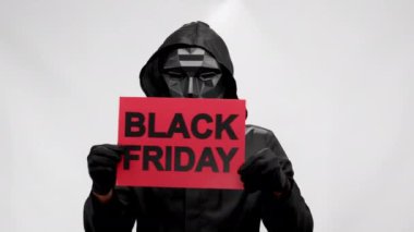 Cheerful anonymous informs about Black Friday showing red plate. Dancing man in Steward mask from Squid Game and black coat with hood invites to take advantage of Black Friday