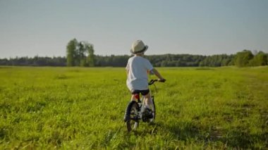 Child boy is riding bike with pinwheel on green field in countryside at summer day, back view. Outdoors activities on nature for kids, freedom and happy childhood concept. Boy in hat on bicycle.