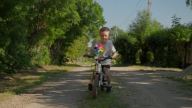 Happy smiling child boy is riding a bike with pinwheel on a country road in slow motion. Kid looks into camera with his tongue sticking out. Outdoors activities for kids and happy childhood concept.