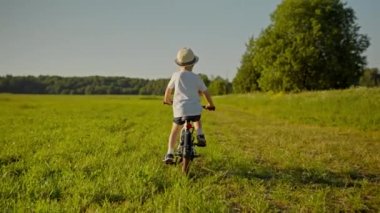 Junior schoolboy dressed in white hat and casual clothes rides two-wheeled bicycle. Boy cycles on grass field against high green trees back view slow motion