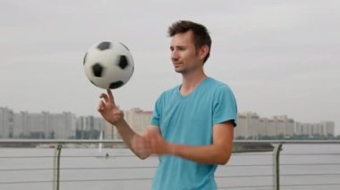 Young man wearing blue t-shirt looks around smiling. Stylish sportsman juggles football on finger. Brown-haired freestyler enjoys hobby against city closeup
