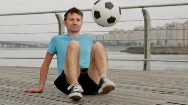 Young freestyler juggles ball sitting on wooden boardwalk. Sportsman demonstrates juggling skills. Stylish man kicks ball using toes against blurry grey buildings
