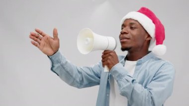 Energetic young African-American guy in Santa hat announces opening of Black Friday via megaphone. Cheerful man speaks actively gesticulating with hand