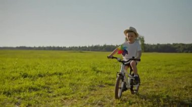 Little child rides white bicycle on grass field. Preschooler in men panama hat and white T-shirt cycles with spinning colorful windmill attached to handlebar slow motion